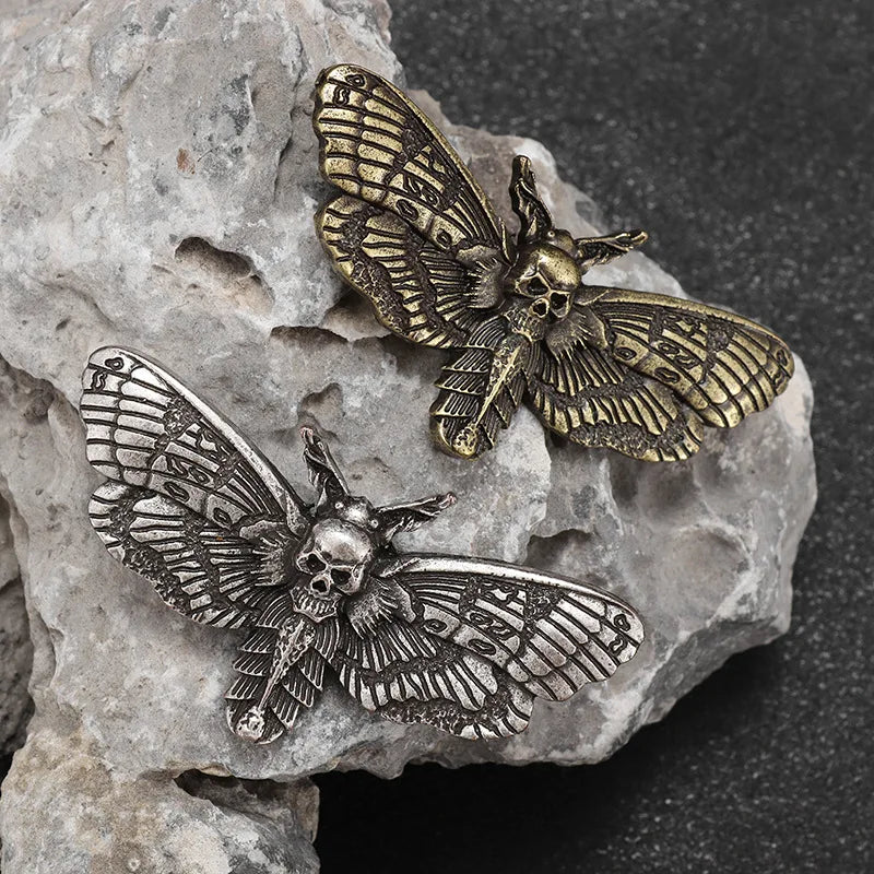 Silver-tone and gold-tone gothic moth brooches displayed on pale textured stone, with dark recesses highlighting their wing patterns.