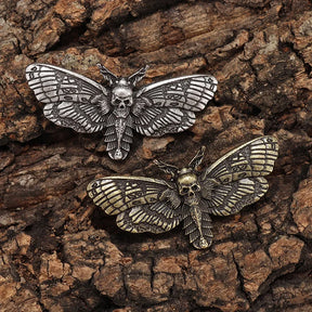 Two death head moth pins arranged on bark, comparing their silver-tone and gold-tone finishes.