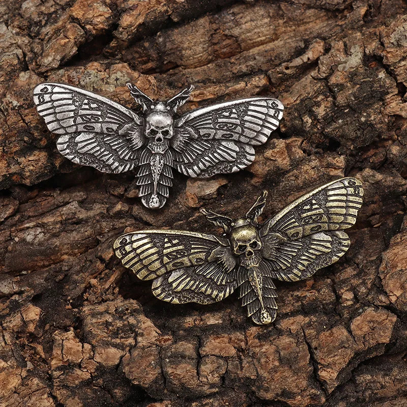 Two death head moth pins arranged on bark, comparing their silver-tone and gold-tone finishes.