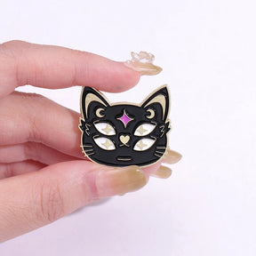 Four-eyed black cat face enamel pin held between fingers, showing pale star-shaped pupils, crescent moons and a purple forehead accent.