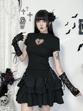 Full outfit view of the black gothic heart-cutout T-shirt with a ruffled skirt and lace arm warmers against a wall decorated with bats and crosses.