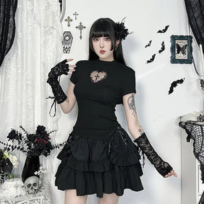 Black heart-cutout T-shirt styled with a short tiered skirt and lace arm warmers in a room with bat and cross decorations.