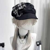 Black gothic newsboy cap with a monochrome plaid patch, safety-pin accents, draped chains and a curved brim.