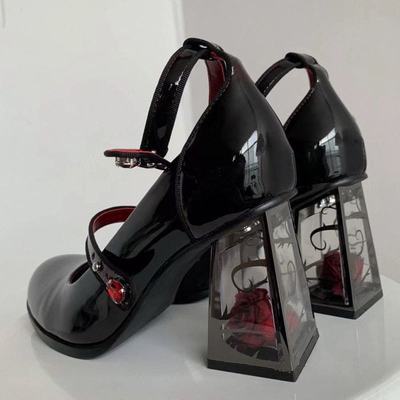 A pair of glossy dark Mary Janes arranged against a pale background, with one shoe turned to show the red rose enclosed in its transparent heel.