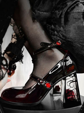 Close side view of a glossy dark wine-red Mary Jane shoe with two slim straps and red jewel accents. A red rose sits inside the clear block heel behind black ornamental lines.