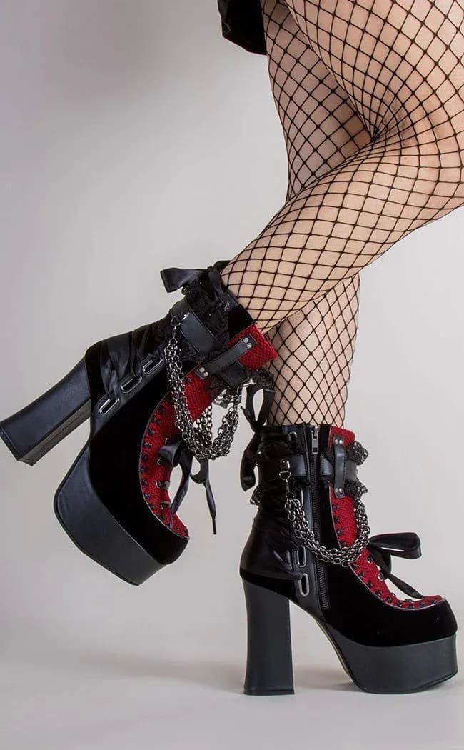 Red gothic ankle boots on crossed legs, showing the platform soles, side zipper and black ribbon bows.