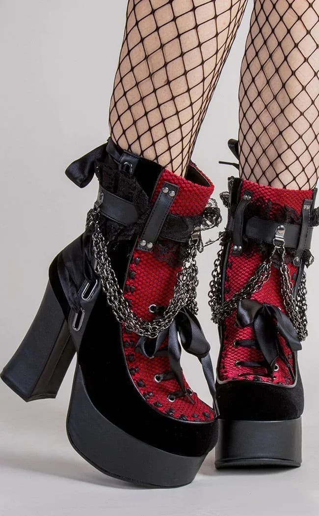 Red gothic platform boots worn with crossed ankles, showing lace panels, black ribbon bows and chunky heels.