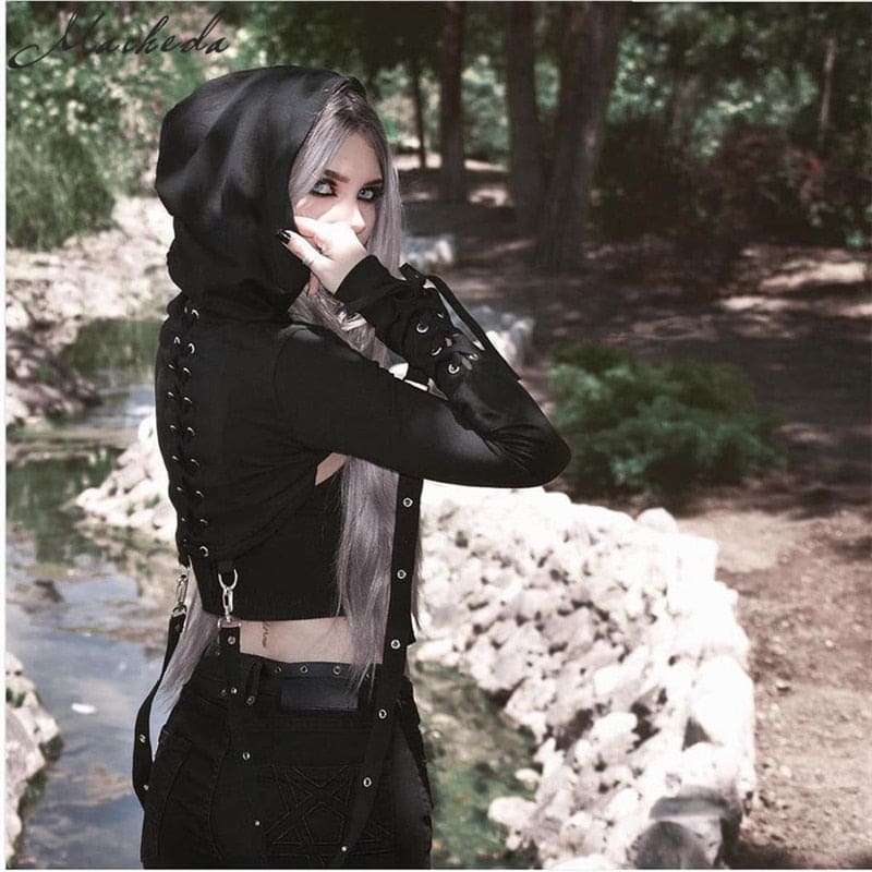 Angled portrait of the black hooded shrug outdoors, showing the raised hood, forearm lacing and a hanging strap.