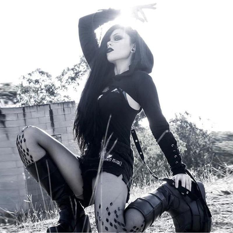 Black hooded crop shrug worn seated outdoors over a separate dark outfit, with lace-up forearms and long hanging straps.