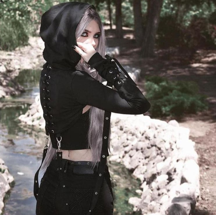 Side view of the black hooded shrug worn in a wooded setting, showing the lace-up forearm and cropped back.