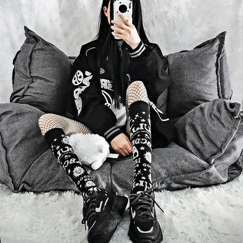 Black-and-white Ouija knee stockings styled with fishnet tights, black shoes and a dark jacket while seated on a grey sofa.