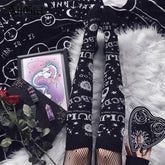 Black Ouija stockings worn on extended legs over fishnet tights, surrounded by a celestial blanket, illustrated books and a planchette-shaped accessory.