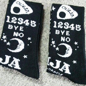 Folded black stockings showing white planchettes, numbers, stars, crescent moons and Ouija lettering on a pale textured surface.