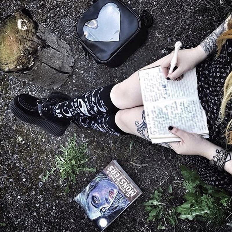 Ouija stockings styled with black shoes and a short dark outfit outdoors, beside an open handwritten book and a heart-shaped bag.