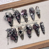Purple and black almond press-on nails arranged on a pale fabric-lined wooden tray, with raised roses, butterfly wings, faceted accents and fine silver-tone chains.