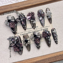 Purple and black almond press-on nails arranged on a pale fabric-lined wooden tray, with raised roses, butterfly wings, faceted accents and fine silver-tone chains.
