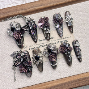 Purple and black almond press-on nails arranged on a pale fabric-lined wooden tray, with raised roses, butterfly wings, faceted accents and fine silver-tone chains.
