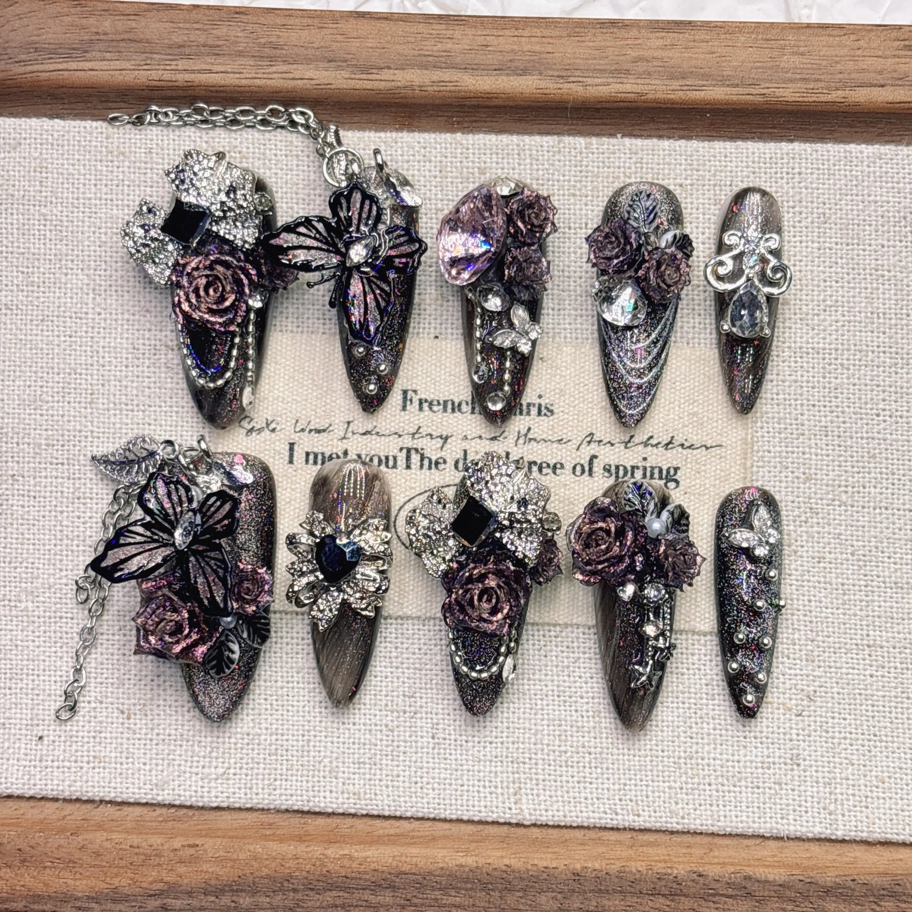 Ten ornate purple gothic press-on nails in two rows, with black heart accents, sparkling stones, dimensional flowers and butterflies against pale fabric.