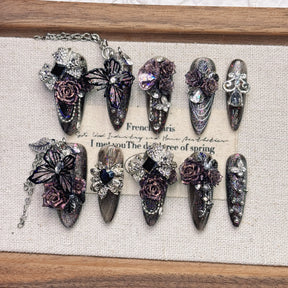 Ten ornate purple gothic press-on nails in two rows, with black heart accents, sparkling stones, dimensional flowers and butterflies against pale fabric.