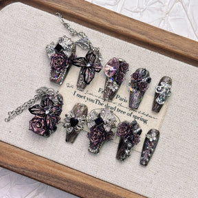 Gothic stiletto press-on nails displayed at an angle on a wooden tray, showing pointed black-purple tips, sculpted roses, butterfly accents and hanging chains.
