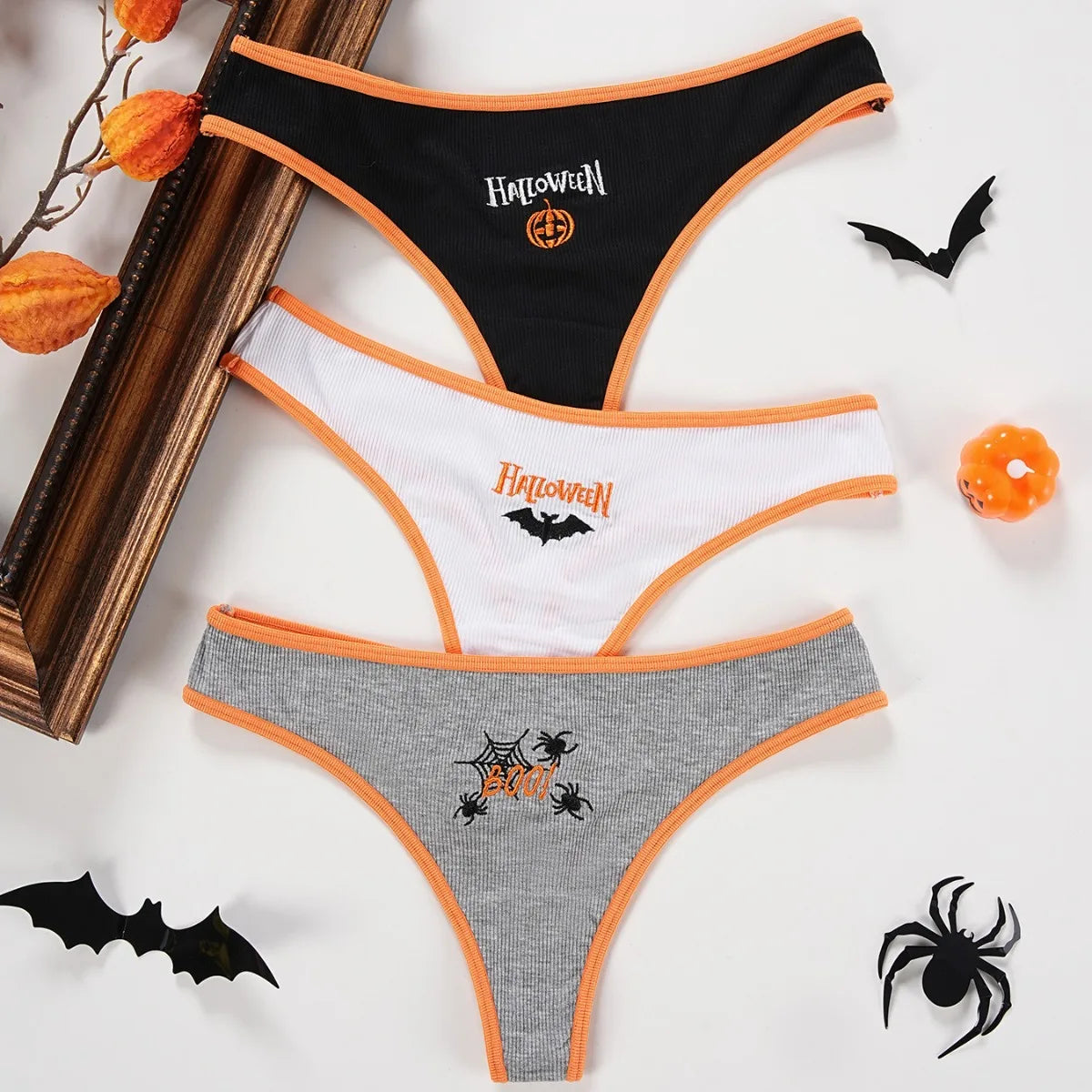 Three Halloween thongs laid flat in black, white and gray, with orange edging and distinct pumpkin, bat and spider motifs.