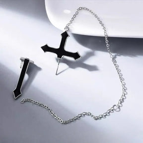 Black cross earring components on a pale surface, showing the piercing post on the upper section and the separate lower section connected by a fine chain.