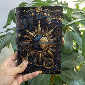 Sun and moon journal held in front of foliage, with a gold-coloured sunburst, crescent face and wrap cord across the dark cover.