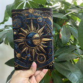 Front view of the dark celestial notebook, with ornate sun, moon and orbital artwork above and below its wraparound cord.