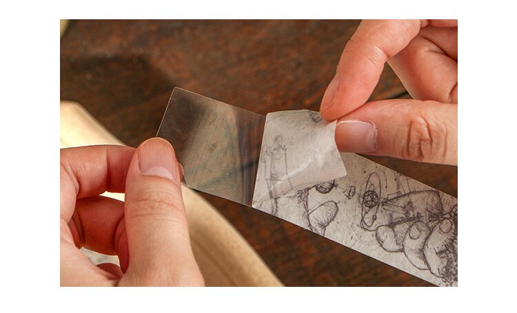 Hands peel the backing from a translucent-looking printed sticker strip with a fine vintage illustration.