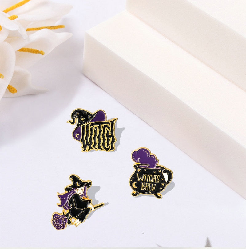 Three black and purple enamel pins show a pointed witch hat, a Witches Brew cauldron and a witch riding a broom beside white display blocks.