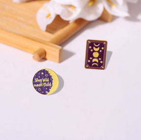 The purple moon-phase card pin and round Stay Wild Moon Child pin displayed together beside a wooden tray and pale flowers.