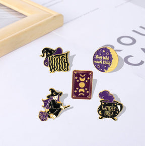 Five witch-themed enamel pins on a pale surface beside a small tray, showing their different silhouettes and coordinated purple accents.