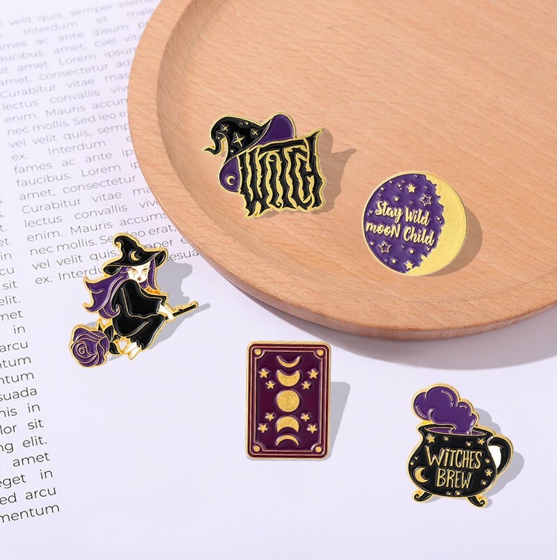 Purple and gold-tone witchery pins displayed beside a wooden tray and a page of text, showing all five designs together.