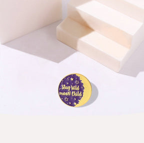 Round purple Stay Wild Moon Child enamel pin with a gold-tone crescent edge, photographed beside pale stepped display blocks.