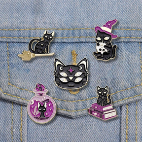 Five witchy black cat enamel pin designs arranged on a blue denim pocket: a broom cat, witch-hat cat with a skull, four-eyed cat face, potion-bottle cat and spellbook cat.