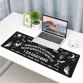 Ouija mousepad beneath a laptop and mouse on a white desk, with a small plant and cup nearby. The black mat features white skeletons, moths, palmistry-style hands and celestial artwork.