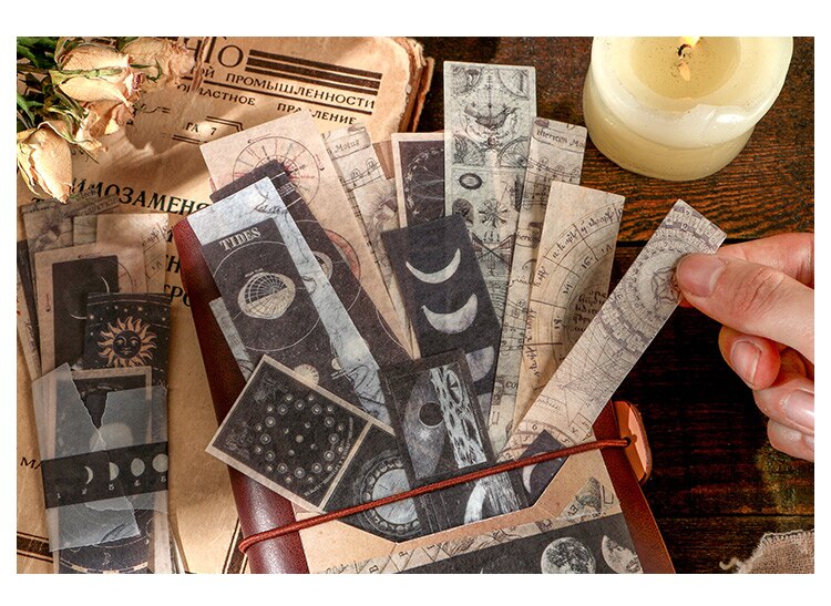Moon-phase and celestial sticker strips tucked inside a brown journal, with a hand holding one strip beside a candle.