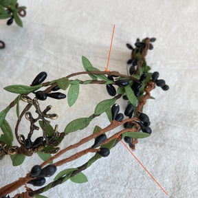 Close-up of brown wrapped branches, green fabric leaves and glossy dark buds on a woodland tiara, with red arrows marking the branch construction.