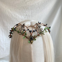 Front view of a leafy antler tiara with dark berries and a black round centre, displayed across pale hair.