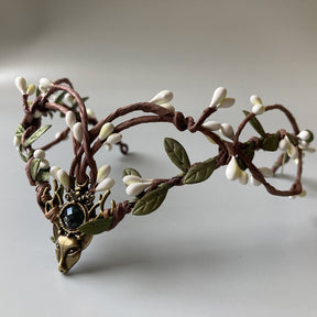 Front view of a woodland tiara with looped brown branches, white buds and a black jewel-like accent above a deer-head motif.