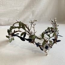 Silver-tone antler tiara with dark buds, green leaves and a black round accent above a small deer-head detail.
