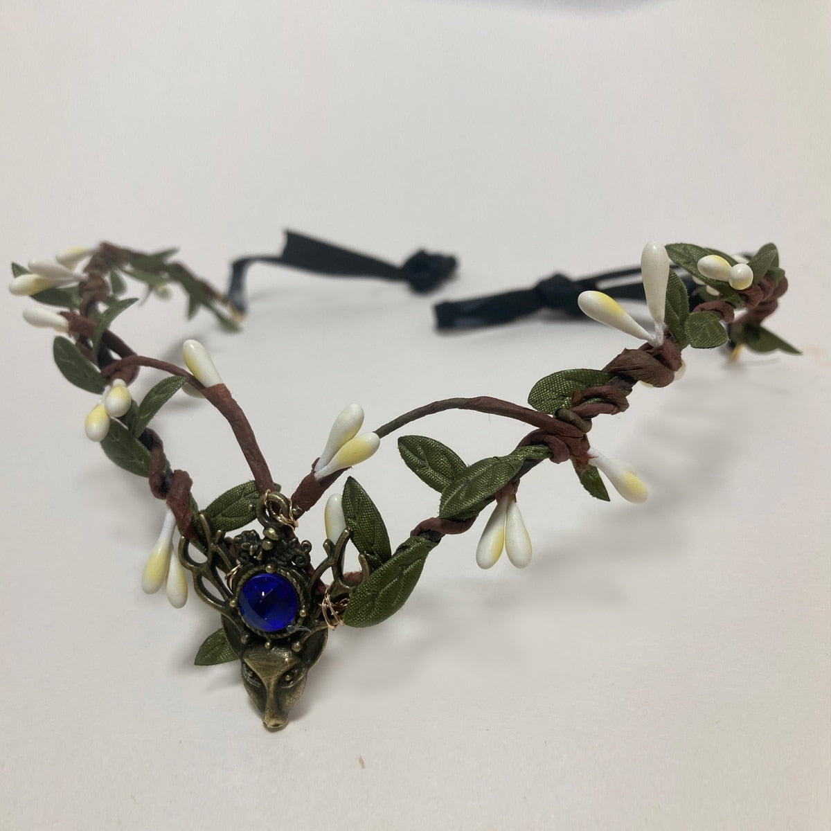 Woodland tiara with pale buds, green leaves and a deep blue round accent above an antiqued deer-head motif.