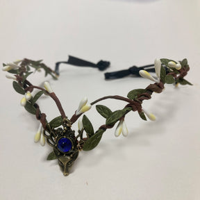 Woodland tiara with pale buds, green leaves and a deep blue round accent above an antiqued deer-head motif.