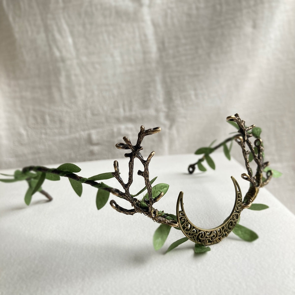 Bronze-tone crescent tiara with green leaves and branching antlers, arranged on pale fabric.
