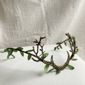 Bronze-tone crescent tiara with green leaves and branching antlers, arranged on pale fabric.