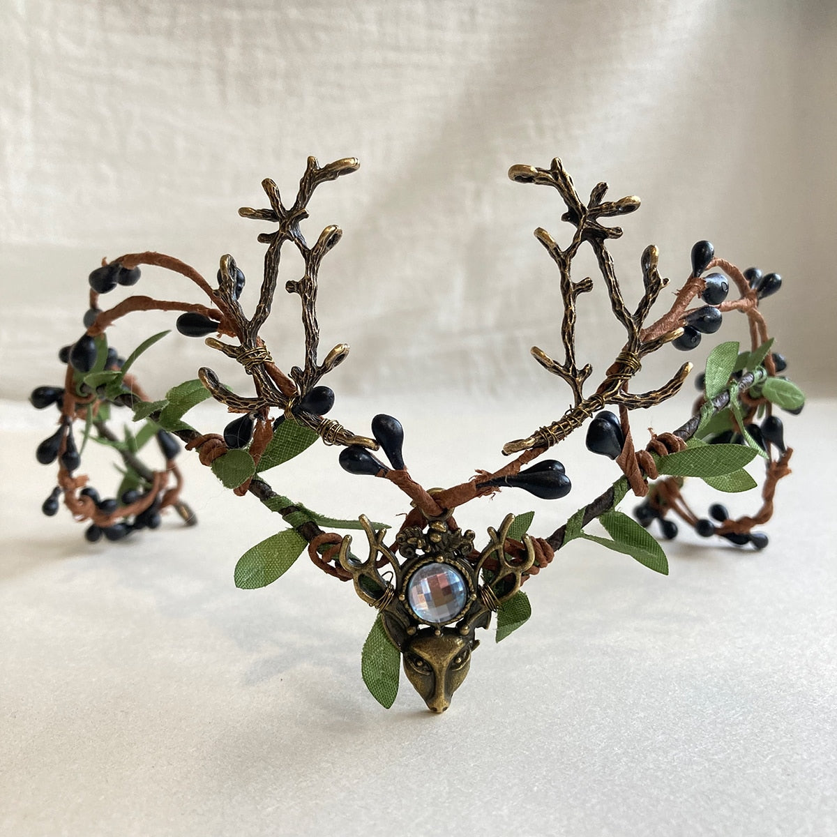 A clear round accent sits above the deer-head motif on a leafy antler tiara with dark berry-like buds.