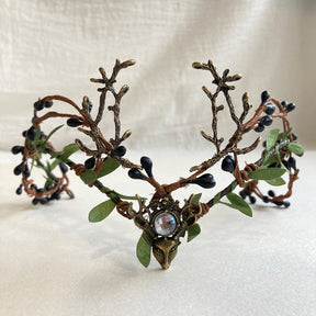 A clear round accent sits above the deer-head motif on a leafy antler tiara with dark berry-like buds.
