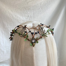 Clear-centred woodland antler tiara displayed on pale hair, surrounded by green leaves and dark berry-like buds.