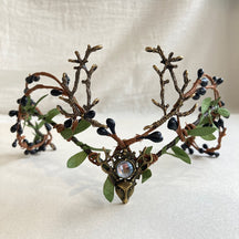 Bronze-tone antler tiara with dark berry-like buds, green leaves and a clear central accent above a deer-head motif.