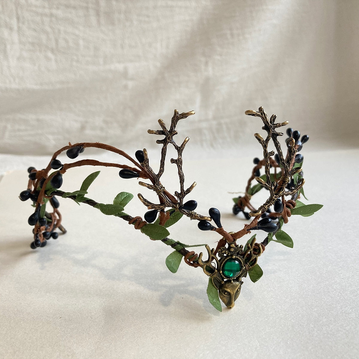 Front view of a bronze-tone woodland antler tiara with a green centre accent and dark buds threaded through leafy branches.